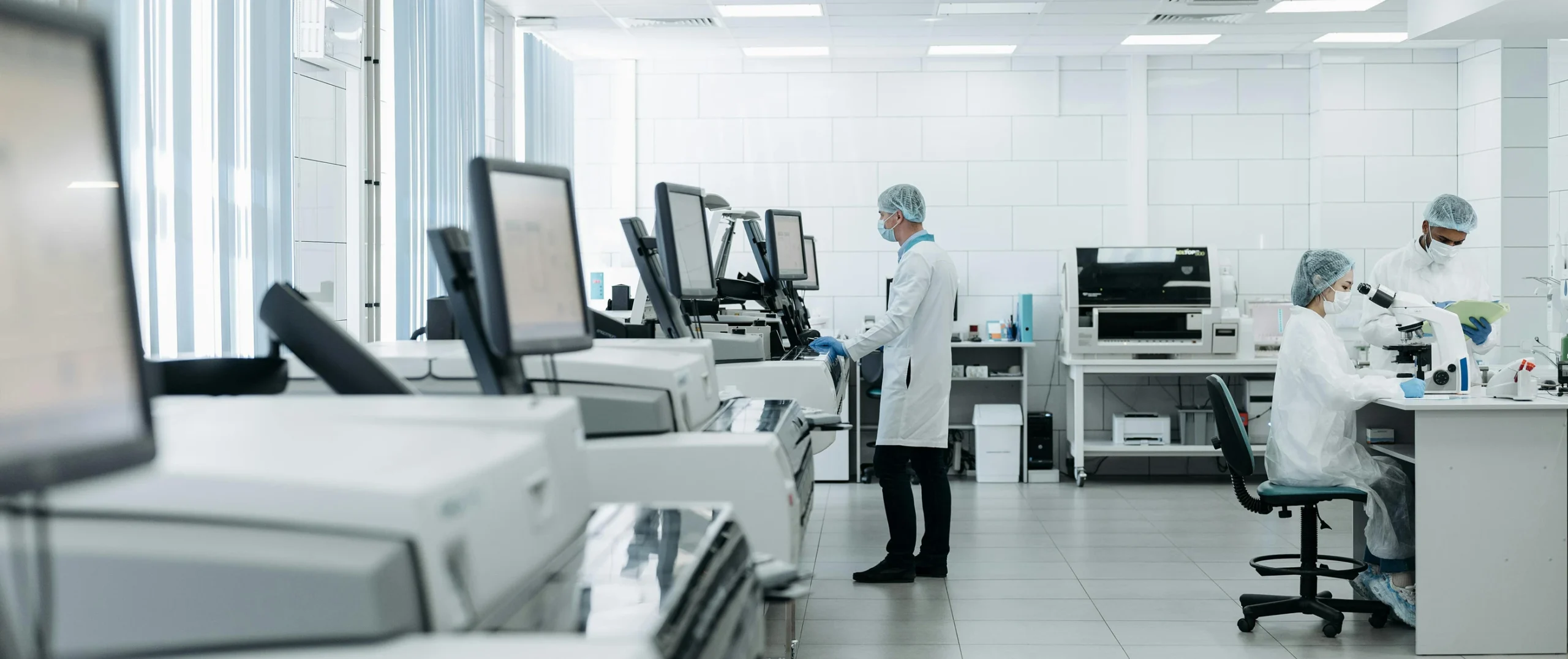 Laboratory with multiple devices, with screens and a worker next to them, and 2 other workers on the right with a microscope and documents.